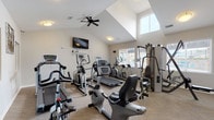 This single-floor fitness and amenity facility is designed to support a well-rounded exercise routine in a bright, comfortable environment. The main fitness area features vaulted ceilings that lend a sense of openness, complemented by a ceiling fan that promotes air circulation during workouts. Natural light streams in through multiple windows with blinds, including elevated clerestory windows that illuminate the space from above, creating an inviting and energizing atmosphere. The walls are finished in a clean, neutral tone, and the floor is covered in low-pile carpet suited for fitness use. Equipment includes recumbent and upright stationary bikes, multiple elliptical cross-trainers, treadmills, and a multi-station cable strength machine, providing options for both cardio and resistance training. A wall-mounted flat-screen television adds entertainment convenience. An interior pass-through window connects the fitness room to an adjacent area, adding a sense of connectivity throughout the facility. A separate laundry room is also part of the space, featuring vinyl plank flooring in a mixed light and dark wood pattern, top-loading washing machines, and a built-in folding shelf — offering a practical and functional complement to the fitness amenities. Fitness room rules are posted on the wall, reflecting an organized and community-oriented environment.