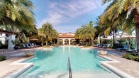 The centerpiece of this outdoor amenity space is a generously sized swimming pool with crystal-clear turquoise water, designed to evoke a true resort atmosphere. The pool stretches expansively, with wide sun shelves on either side providing shallow lounge areas ideal for relaxation. Surrounding the pool, a well-maintained deck is lined with cushioned chaise lounges and shaded by large umbrellas, offering ample space for sunbathing and leisure.

Tall, mature palm trees frame the entire area, creating a lush tropical canopy that enhances the sense of seclusion and tranquility. At the far end of the pool, a Mediterranean-style clubhouse serves as a striking architectural focal point, featuring white stucco walls, graceful arched colonnades, and a warm terracotta tile roof. The structure's symmetrical design adds a sense of elegance and permanence to the setting.

The overall ambiance is bright and open, with abundant natural light reflecting off the water and the surrounding greenery contributing to a vibrant, resort-like environment. The thoughtful landscaping and cohesive architectural design combine to create an inviting and visually appealing outdoor retreat.