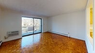This single-floor residence is defined by its warm, glossy parquet wood flooring, which extends consistently throughout the living areas and bedroom, lending a cohesive and timeless character to the space. The living room is generously proportioned, featuring a large sliding glass door that opens onto a balcony, flooding the room with natural light and providing outdoor views of surrounding greenery and neighboring buildings. A baseboard heating system runs along the perimeter walls, and a wall-mounted HVAC unit offers additional climate control.

The kitchen is accessed via a pass-through opening from the living area, and is outfitted with light wood cabinetry, ample counter space, a double sink, an electric range, and a full-size refrigerator. Overhead lighting illuminates the workspace efficiently. The layout is compact yet functional, with vinyl flooring distinguishing it from the rest of the unit.

The bedroom offers a comfortable footprint, a window with outdoor views, and a wide closet with bi-fold doors, providing practical storage. A textured ceiling is present throughout the unit.

The bathroom is fitted with distinctive pink tile walls and a matching tub and shower surround, mosaic tile flooring, a vanity with an undermount sink, and a wall-mounted cabinet above the tub — a retro aesthetic detail that adds character to the space.