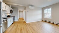 This well-appointed single-floor residential unit offers a clean, modern aesthetic throughout. The open living area is anchored by light-toned hardwood flooring that extends seamlessly through the main living spaces, complemented by neutral gray-white walls and crisp white trim. Multiple windows fitted with white horizontal blinds invite abundant natural light and provide views of the surrounding neighborhood. A wall-mounted mini-split HVAC unit ensures year-round climate comfort alongside the baseboard heating present throughout the unit.

The kitchen is thoughtfully designed with white shaker-style cabinetry, granite countertops featuring subtle gray and white veining, a stainless steel double-basin sink, and a full suite of stainless steel appliances including a dishwasher, range, and over-the-range microwave. The layout flows efficiently into the main living area, making the space ideal for everyday living.

A hallway connects the living areas to the private rooms, where plush neutral carpeting provides a comfortable, quiet atmosphere. The bedroom benefits from corner window placement, enhancing natural light. The bathroom is accessible from the hallway and features granite-topped vanity cabinetry consistent with the kitchen's finishes. Throughout the unit, the consistent use of light materials and neutral tones creates a cohesive, inviting ambiance.