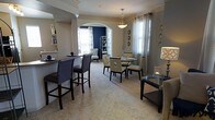 This single-floor space showcases a refined open-concept layout that seamlessly connects the dining and living areas through a graceful arched entryway — a standout architectural detail that adds character and visual depth. The dining area is anchored by a round glass-top table surrounded by cream upholstered chairs, bathed in warm light from a pendant fixture above. Crown molding runs throughout, lending a polished, finished look to each room. A breakfast bar with dark leather counter-height stools creates a casual dining option, thoughtfully positioned between the kitchen and main living space. The soft, neutral carpet adds warmth underfoot and ties the open areas together cohesively. Natural light filters in through multiple windows dressed with sheer curtains, brightening the interior and highlighting the neutral gray wall palette. A bold navy accent wall in the adjacent sitting area provides a striking contrast, adding personality and depth to the overall design. Decorative wall accents, a side table with a stylish lamp, and built-in shelving complete the curated aesthetic. The space strikes a balance between comfort and sophistication, making it both functional and visually inviting.