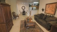 This single-floor space showcases an open-concept design that seamlessly connects the living, dining, and kitchen areas into one cohesive environment. The living area is anchored by a plush, dark upholstered sofa paired with a substantial wood coffee table resting atop a traditional patterned area rug, creating a warm and inviting focal point. An ornate accent chair adds a classical touch near the entryway corridor, complemented by a decorative wall sconce and a large round wall clock that adds character to the neutral-toned walls. The dining area, positioned mid-space, features an elegant wrought-iron and wood table set with upholstered chairs, suitable for intimate gatherings. Beyond the dining area, the kitchen is visible and equipped with light wood cabinetry, a white refrigerator, microwave, and a raised breakfast bar that provides a natural division between cooking and living spaces. Overhead, recessed and fluorescent lighting ensures the space is well-lit throughout. The consistent use of beige carpeting and warm neutral wall tones ties all areas together, producing a comfortable, classic ambiance.