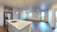 This single-floor unit offers a thoughtfully designed open-concept layout with consistent use of wood-look flooring throughout the main living areas. The kitchen serves as a central feature, equipped with grey cabinetry extending to the ceiling, a white quartz island with an undermount sink, subway tile backsplash, stainless steel appliances including a French door refrigerator and electric range, and a dedicated microwave. Three pendant lights hang above the island, providing focused task lighting. The kitchen flows seamlessly into a spacious living area, which is brightened by a large window and a glass door leading to a private balcony with open outdoor views, including distant mountain scenery. A ceiling fan serves the living space.

The bedrooms feature plush carpeting and ceiling fans, with large windows framing expansive mountain and landscape views. The primary bathroom offers a dual-sink vanity with quartz countertops, grey cabinetry, a large mirror, and a separate toilet area, while a secondary bathroom includes a single-sink vanity and a full tub-shower combination. A dedicated utility room houses a full-size washer and dryer alongside the HVAC and water heater systems.