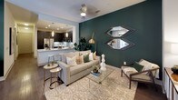 This well-appointed single-floor residence showcases a thoughtfully designed open-concept layout connecting the kitchen, dining, and living areas. The kitchen is a standout feature, offering dark cabinetry with glass-front upper cabinets, a light quartz peninsula with seating for multiple bar stools, a subway tile backsplash, stainless steel appliances, and pendant lighting. A bold teal accent wall anchors the living area, which flows seamlessly into the kitchen and is finished with dark hardwood-style flooring. The dining area is positioned near large windows with floor-to-ceiling curtains, allowing natural light to fill the space, alongside built-in cabinetry ideal for storage and display. Two bedrooms feature plush carpeting; one includes a dark slate accent wall with a gallery arrangement, while the other offers neutral tones and natural light from a large window. Both bathrooms are finished with dark vanity cabinetry, gray countertops, large mirrors, tile flooring, and shower-tub combinations. The cohesive use of dark cabinetry, teal and slate accents, and warm gold hardware creates a contemporary and polished aesthetic throughout.