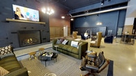 This sophisticated lounge space makes a strong first impression with its bold, industrial-chic design. The interior is anchored by a striking floor-to-ceiling fireplace clad in dark tile with a herringbone pattern, providing a warm focal point for the main seating area. Deep navy and charcoal walls, paired with warm wood wainscoting panels, create a rich, layered atmosphere throughout the space.

The open-plan layout is furnished with a curated mix of seating arrangements, including a plush olive-green channeled sofa, leather club chairs, and high-back accent chairs in tan and navy — offering a variety of comfortable gathering spots. Houndstooth-patterned area rugs define the seating zones and add a refined, textural contrast to the overall design.

Overhead, exposed metal ductwork and industrial-style conduit run along the dark ceiling, lending an urban edge to the space. A combination of statement globe pendant lights and recessed fixtures provide layered illumination, enhancing the lounge's inviting ambiance.

Wood-paneled walls with wainscoting detailing add a classic architectural element that balances the industrial components. The generous ceiling height amplifies the sense of openness, making this a well-appointed and versatile communal space ideal for relaxing or socializing.