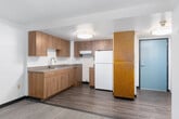 This single-floor unit is thoughtfully laid out to include a main living area, a separate room, a kitchenette, and a full bathroom. The living and sleeping areas are finished with patterned carpet in muted blue-gray tones, complemented by neutral off-white walls and dark baseboards. Textured popcorn ceilings run throughout, and baseboard heating units are installed beneath the windows in each carpeted room. One room features a painted concrete block accent wall, lending a durable, institutional character to the space. Windows in both carpeted rooms frame views of lush, mature trees, bringing in natural light and a connection to the outdoors.

The kitchenette is outfitted with warm wood-toned cabinetry, ample upper and lower storage, gray composite countertops, a stainless steel sink, and a range hood. Wood-look vinyl plank flooring extends through the kitchen and hallway areas, creating a practical transition between spaces. A closet with louvered doors is accessible from the entry hall.

The bathroom is designed with accessibility in mind, featuring multiple stainless steel grab bars near the toilet and bathtub, a dark-cabinet vanity with a mirror, and wood-look vinyl flooring, providing both functionality and safety.