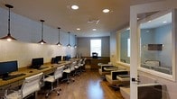 This single-floor space is designed to serve as a functional and inviting business center or shared workspace. The layout is thoughtfully organized, combining a dedicated computer workstation area with a comfortable lounge seating section. Along one wall, a long wooden desk surfaces multiple desktop computer setups, each accompanied by ergonomic white rolling chairs. A row of industrial-style black pendant lights hangs above this work area, providing focused task lighting that complements the recessed ceiling lights overhead. The warm-toned hardwood flooring runs throughout the space, lending a cohesive and polished look. Natural light filters in through windows along the far wall, brightening the neutral gray walls and creating an open, airy atmosphere. A built-in wooden cabinet and shelving unit near the windows adds additional storage and a seamless architectural detail. On the opposite side of the room, a lounge area features modern square chairs with cream upholstery and dark frames, offering a relaxed setting for informal meetings or casual use. The overall ambiance strikes a balance between professional functionality and comfortable design, making this space well-suited for productivity and collaboration.