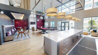 This expansive single-floor amenity space is designed with a contemporary aesthetic and an open, airy layout that encourages social interaction. The centerpiece of the space is a dramatic double-height atrium flooded with natural light through large floor-to-ceiling windows and glass doors that offer views of the surrounding courtyard. Striking drum pendant lights and mirrored globe fixtures hang from the soaring ceiling, adding a sophisticated visual focal point overhead.

The main lounge area features multiple seating arrangements, including modern accent chairs, leather lounge seating, and patterned area rugs that define individual gathering zones. A bold, vibrant mural serves as a dramatic accent wall near the kitchen, which includes a large white quartz island, built-in appliances, and bar-height seating along an extended counter — ideal for casual dining or socializing.

A full-size pool table anchors the game zone, complemented by booth-style seating with metallic upholstery set against decorative branch-patterned wall panels. A dedicated reception or concierge area features a clean-lined counter, wall-mounted display, and linear ceiling lighting. Throughout, warm wood-tone flooring, dark structural accents, and rich burgundy wall panels create a cohesive, upscale atmosphere that balances energy and comfort.