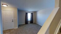 This two-floor space presents a straightforward and functional layout with clean, neutral interiors throughout. The pictured room features wall-to-wall carpeting in a muted gray-beige tone, providing a soft underfoot feel that complements the light gray walls. A single window dressed with dark charcoal curtains allows natural light to filter into the room, creating a calm and understated ambiance. White baseboards trim the perimeter of the room, adding a crisp contrast to the neutral palette. The textured ceiling adds a subtle visual element to the space. A doorway on the left side of the room suggests access to an adjacent area, possibly a closet or hallway, while the staircase visible in the corner indicates the connection between the two floors of the property. The overall aesthetic is simple and move-in ready, with a neutral color scheme that offers flexibility for a variety of furnishing styles. The space maintains a practical and uncluttered character, with no overly decorative elements, making it well-suited for those seeking a clean and adaptable living environment across its two levels.