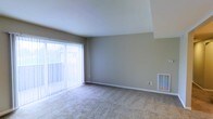 This single-floor space offers a clean, functional layout designed for comfortable everyday living. The main living area is finished with wall-to-wall carpeting in a neutral tone, providing a warm and quiet underfoot surface throughout. Walls are painted in a soft greige hue, contributing to a calm and cohesive aesthetic that pairs well with a variety of furnishing styles.

A standout feature of the living area is the vaulted ceiling, which adds a sense of height and openness, making the space feel larger and more airy than a standard flat ceiling would allow. Natural light is abundantly supplied through a full-height sliding glass door fitted with vertical blinds, offering both privacy control and easy access to an outdoor area. The greenery visible through the glass suggests a pleasant outdoor setting just beyond the unit.

A wall-mounted HVAC vent is positioned at floor level, ensuring efficient climate control throughout the space. White baseboards provide a crisp contrast against the neutral walls and carpet, adding a subtle but polished finishing detail. The overall ambiance of the space is bright, clean, and move-in ready, with a straightforward design that offers a versatile canvas for personalization.