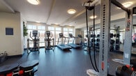 This fitness center is designed to support a full range of workout routines in a bright, well-organized environment. Large windows line the walls, flooding the space with natural light and providing open views to the exterior, creating an energizing atmosphere for exercise. The rubber-floored workout area is thoughtfully divided into distinct training zones. The cardio section is outfitted with multiple treadmills, elliptical trainers, and stationary bikes, all arranged to take advantage of the natural light from the surrounding windows. A dedicated strength training area features a multi-functional cable machine and rack system, allowing for a wide variety of resistance exercises. Complementing these is a functional training zone stocked with free weights, dumbbells, medicine balls, plyo boxes, resistance bands, and a cable-based functional trainer, providing ample options for dynamic and sport-specific training. Full-length mirrors are mounted along the walls, enhancing the sense of space and allowing users to monitor their form throughout their workouts. Flush-mounted ceiling lights provide consistent overhead illumination throughout the facility. The space is finished with a clean, modern aesthetic, featuring neutral-toned walls and a durable, dark rubber floor designed to withstand heavy use. A water station and informational signage are also present, rounding out the amenities of this comprehensive fitness facility.