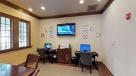 This single-floor business center is designed to meet the productivity needs of its visitors with a clean, professional layout. The room features two dedicated computer workstations, each equipped with a desktop monitor and a comfortable upholstered chair, providing ample space for focused work. A large flat-screen television is mounted prominently on the center wall, capable of displaying news, presentations, or other media content. Between the workstations sits a dark wood cabinet unit that houses a printer, offering convenient document printing access. The walls are finished in a warm neutral tone and accented with decorative chevron-patterned panels and subtle wall art, adding a touch of visual interest without overwhelming the functional atmosphere. Crown molding along the ceiling and rich wood baseboards contribute a refined, polished character to the space. Recessed lighting illuminates the room evenly, ensuring a comfortable environment for extended use. The light-toned flooring ties the room together, creating a bright and inviting setting that balances practicality with thoughtful design.