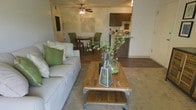 This single-floor residence features an open-concept layout that seamlessly connects the living, dining, and kitchen areas into one cohesive space. The living area is anchored by plush neutral-toned seating complemented by green accent pillows, centered around a rustic wood-and-metal coffee table that adds an industrial charm. A ceiling fan overhead provides both function and a casual aesthetic to the room.

Transitioning naturally from the living space, the dining area accommodates a small table paired with tufted green upholstered chairs, creating an inviting spot for meals. The hardwood-style flooring in this zone adds warmth and continuity, while the carpeted living area provides a soft contrast underfoot.

The kitchen is visible beyond the dining space, featuring dark wood cabinetry and a raised breakfast bar that serves as a natural dividing element between the cooking and dining zones. The overall color scheme throughout the space relies on earthy neutrals and green accents, creating a cohesive and welcoming ambiance. Decorative greenery and glass vessels add organic touches throughout the open layout.