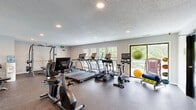 This single-floor fitness center offers a generously sized workout space designed to accommodate a comprehensive range of exercise equipment and activities. The room features durable speckled rubber flooring throughout, providing both safety and functionality for high-impact workouts. Recessed lighting is embedded in the textured ceiling, ensuring consistent, even illumination across the entire space.

The cardio section is well-appointed with multiple True-brand treadmills, ellipticals, and stationary bikes, each equipped with individual display screens. A row of windows along one wall allows abundant natural light to fill the space while offering views of a neighboring outdoor balcony area, enhancing the workout environment with an open, airy feel.

The strength training area includes a multi-station cable machine, a dumbbell rack, and additional resistance equipment, providing users with diverse options for full-body conditioning. Accessories such as medicine balls, exercise mats, and stability balls are also available. A dedicated corner houses a workstation with a desk, and restroom facilities are accessible directly from the fitness floor via dark-framed doors. The neutral gray walls and clean layout contribute to a practical, organized, and motivating atmosphere throughout.