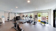This single-floor fitness center offers a generously sized workout space designed to accommodate a comprehensive range of exercise equipment and activities. The room features durable speckled rubber flooring throughout, providing both safety and functionality for high-impact workouts. Recessed lighting is embedded in the textured ceiling, ensuring consistent, even illumination across the entire space.

The cardio section is well-appointed with multiple True-brand treadmills, ellipticals, and stationary bikes, each equipped with individual display screens. A row of windows along one wall allows abundant natural light to fill the space while offering views of a neighboring outdoor balcony area, enhancing the workout environment with an open, airy feel.

The strength training area includes a multi-station cable machine, a dumbbell rack, and additional resistance equipment, providing users with diverse options for full-body conditioning. Accessories such as medicine balls, exercise mats, and stability balls are also available. A dedicated corner houses a workstation with a desk, and restroom facilities are accessible directly from the fitness floor via dark-framed doors. The neutral gray walls and clean layout contribute to a practical, organized, and motivating atmosphere throughout.