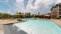 Located at 3500 Carrington Mill Blvd in Morrisville, NC, this outdoor amenity space is defined by its impressive resort-style pool area, designed to offer a premium leisure experience. The generously sized swimming pool features a freeform shape with multiple entry points, water features, and a dedicated wading section, all framed by a spacious stamped concrete deck. Bright yellow market umbrellas and rows of lounge chairs line the pool's perimeter, creating a vibrant and inviting atmosphere perfect for relaxation.

Wooden pergolas are strategically positioned throughout the space, providing shaded gathering areas furnished with seating for outdoor entertaining. A built-in outdoor kitchen station, complete with a stainless steel gas grill set into a stone surround, adds excellent functionality for alfresco dining. The surrounding stone and brick architectural facade of the multi-story building lends the area a refined, upscale character.

The space is bordered by mature trees that provide a natural, verdant backdrop and a sense of privacy. A dedicated clubhouse entrance, featuring large glass doors framed in brick with covered porch seating, seamlessly connects the indoor amenities to the outdoor environment. The abundant natural light and open-sky setting enhance the overall resort-like ambiance of this well-appointed outdoor retreat.