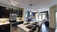This contemporary single-floor space showcases a thoughtfully designed open-concept layout that seamlessly connects the kitchen, dining, and living areas. The kitchen is a standout feature, outfitted with dark espresso cabinetry — including glass-front upper cabinets — stainless steel appliances such as a range, oven, and built-in microwave, and a sleek subway tile backsplash. The centerpiece of the kitchen is a large island with a crisp white countertop, an undermount double sink, and bar-height seating, making it ideal for both casual dining and entertaining. Track lighting and elegant pendant lights illuminate the space with a warm, modern ambiance. The open living area beyond benefits from abundant natural light streaming through large floor-to-ceiling windows, offering views to the outdoors. A bold navy blue accent wall adds a striking visual anchor to the living space, which is complemented by wood-tone flooring throughout. A ceiling fan overhead enhances comfort in the living area. The overall design balances bold contrasts — dark cabinetry against white surfaces, rich color accents against neutral tones — creating a sophisticated and inviting atmosphere throughout.
