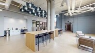 This single-floor commercial workspace masterfully blends industrial architecture with contemporary design. Soaring ceilings with exposed ductwork, structural concrete columns, and polished concrete floors establish a loft-like aesthetic throughout the open main area. A striking geometric pendant light installation anchors the central co-working zone, where light wood high-top communal tables paired with sleek black bar stools provide versatile workspaces for individuals or groups.

A lounge area offers a more relaxed setting, furnished with a neutral-toned sofa, marble-top coffee tables, and accent chairs, all bathed in abundant natural light streaming through oversized, floor-to-ceiling industrial-style windows. Adjacent to the lounge, a kitchenette features white shaker cabinetry, a matte black faucet with an undermount sink, light quartz countertops, and a white subway tile backsplash, along with a built-in beverage refrigerator.

Private offices enclosed by full-height glass walls and dark-framed partitions offer focused work environments while maintaining visual connectivity to the broader space. A metal staircase with rail accents and an elevator provide access between levels. The overall ambiance is polished yet approachable, balancing open collaboration with private functionality in a refined industrial setting.