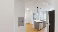 Unit 607 at 231 E Main St in Bound Brook, NJ offers a efficiently designed one-bedroom, one-bathroom layout on a single floor. The open-concept living and kitchen area anchors the unit, featuring a white kitchen island with an undermount sink, sleek gray cabinetry extending to the ceiling, and a full suite of stainless steel appliances including a refrigerator, electric range, and built-in microwave. Pendant lighting and track lighting illuminate the kitchen, complementing the natural light that enters through the window. Warm-toned hardwood-style flooring runs consistently throughout the unit, lending a cohesive and clean aesthetic.

The spacious bedroom accommodates the majority of the unit's footprint, offering a bright and airy feel thanks to a large window with horizontal blinds that frame exterior views. A wall-mounted PTAC unit provides individual climate control. The neutral gray walls throughout the unit create a calm, modern atmosphere.

The bathroom is finished with large-format light gray wall tiles and features a glass-enclosed walk-in shower with chrome fixtures, conveying a polished, contemporary feel. White panel doors and clean white trim complete the refined interior throughout the unit.