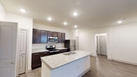 This well-appointed single-floor unit offers a thoughtfully designed open-concept layout that seamlessly connects the kitchen and living areas. The kitchen serves as the centerpiece, featuring a large central island with a granite countertop and undermount sink, rich dark espresso cabinetry with modern hardware, and a full suite of stainless steel appliances including a refrigerator, electric range, and built-in microwave. Recessed lighting illuminates the space, enhancing the clean, contemporary aesthetic throughout.

The living area extends naturally from the kitchen and is finished with warm, wood-style laminate flooring and a ceiling fan, creating a comfortable and functional space. Multiple windows allow natural light to filter in, adding brightness to the neutral-toned walls.

A dedicated bedroom area includes a walk-in closet with built-in shelving, offering generous storage. The bathroom is finished with matching granite countertops, an undermount sink, and a large wall mirror with overhead lighting, maintaining a consistent design language throughout the unit.

Light-toned laminate flooring flows through the bedrooms and hallways, while crisp white trim and panel-style doors complete the polished interior. Overall, this space combines modern finishes with a practical, open layout designed for everyday comfort.