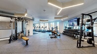 This well-appointed fitness center offers a comprehensive exercise environment designed to support a full range of workout routines. The space is organized into distinct functional zones, beginning with a generous cardio area lined with treadmills, ellipticals, and rowing machines, all positioned to take full advantage of large, floor-to-ceiling windows that flood the room with natural light and provide open outdoor views. The strength training section is equally well-equipped, featuring a full rack of dumbbells, adjustable benches, a cable machine, a power rack with barbell, and additional resistance machines to support both targeted and compound movements. A dedicated free-weight and functional training area provides open floor space for varied exercises. The interior design is modern and clean, with crisp white walls, high ceilings, and a standout geometric LED lighting system that adds a contemporary architectural accent overhead. Full-length mirrors along the walls enhance the sense of space and support form-monitoring during workouts. Durable rubber-tile flooring covers the entire area, offering both safety and noise reduction. Wall-mounted flat-screen TVs are positioned throughout for entertainment. The overall ambiance is bright, professional, and energizing, making this fitness center a fully equipped destination for all levels of fitness activity.
