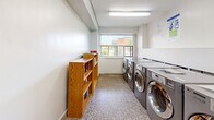Located at 24 Ida Street in St. Catharines, Ontario, this shared laundry facility comprises two dedicated laundry rooms across a single floor, offering a practical and well-organized space for residents. The facility is finished with durable terrazzo-style flooring throughout, bright white walls, and overhead fluorescent lighting that ensures a clean, well-lit environment.

The first laundry room is a larger, more comprehensively equipped space featuring a row of commercial-grade front-loading LG washers and dryers, a stainless steel utility sink with mounted hardware, and a stainless steel folding table for added convenience. Informational signage and notices are posted on the walls, and two grey access doors — one single and one double — provide entry and exit points. A security camera is also present, contributing to a safe environment.

The second laundry room offers a similarly functional layout, with a row of front-loading washers and dryers lining one wall. A window allows natural light to filter into the space, creating a more open atmosphere. A wooden bookshelf with reading materials adds a welcoming touch, while a seating chair provides a comfortable spot for residents to wait. Both rooms are designed with practicality and ease of use in mind, making this facility a valuable shared amenity.