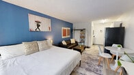 Located at 1057 Broward Rd in Jacksonville, Florida, this efficiently designed single-floor unit offers a well-organized layout comprising one bedroom and one bathroom. The main living area doubles as the bedroom, characterized by a striking deep-blue accent wall that serves as an eye-catching focal point against the surrounding neutral white walls. Light wood-style flooring runs throughout, lending warmth and continuity to the space.

A large floor-to-ceiling window with vertical blinds dominates one wall, allowing generous natural light to fill the room while offering views of surrounding greenery. The space accommodates both sleeping and living functions, with designated zones for a bed, a seating area with a tufted leather sofa and coffee table, an open shelving unit, and a dedicated workspace featuring a desk and wall-mounted television.

The bathroom is accessed through a white paneled door and features marble-look tile on both the floors and tub surround, creating a clean and polished aesthetic. A combination shower and bathtub with a curtain rod, a toilet, and a wall-mounted vanity with mirror complete the bathroom. The textured ceiling throughout adds a subtle classic touch. Overall, the unit is compact yet functional, making efficient use of every square meter for comfortable day-to-day living.