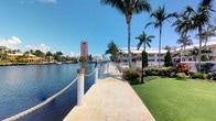 This waterfront property offers a striking blend of tropical charm and urban convenience, set along the edge of a calm, scenic canal. The exterior showcases a beautifully paved stone promenade that runs parallel to the water, finished in warm beige tones and bordered by elegant rope-and-post railings that lend a nautical character to the space. Towering palm trees line the walkway, providing shade and reinforcing the lush, tropical atmosphere. The well-maintained grounds feature vibrant flowering shrubs and manicured green lawn areas, creating a polished and inviting outdoor environment. From the promenade, sweeping waterway views stretch outward, with a notable high-rise tower and additional low-rise buildings visible across the canal, offering a sense of the broader urban context. A pedestrian bridge is visible in the mid-distance, connecting to the surrounding waterfront district. The property benefits from abundant natural light, open skies, and a refreshing outdoor ambiance that is characteristic of South Florida waterfront living. Overall, the space presents a well-appointed, single-level outdoor setting that combines natural beauty with thoughtfully designed hardscape elements, making it an appealing and functional waterfront destination.