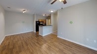 This single-floor unit offers a functional and well-organized layout across several distinct living spaces. The open-concept living and dining area is finished with warm hardwood-style flooring and neutral gray walls, creating a versatile backdrop for any furnishing style. A ceiling fan overhead and a paneled entry door anchor the space, which flows naturally toward the kitchen. A textured ceiling adds subtle character to the living area.

The galley-style kitchen features warm wood cabinetry with dark hardware, dark laminate countertops, and hardwood-style flooring that continues from the main living area. Black appliances, including a refrigerator and range, are integrated alongside a peninsula that provides additional prep space and separation from the living area. A dedicated in-unit laundry area is accessible directly from the kitchen, housing a full-sized washer and dryer with overhead shelving for added storage convenience.

The bedrooms are finished with plush carpeting and neutral beige walls, offering a comfortable and quiet retreat. Natural light enters through a window fitted with horizontal blinds. The bathroom presents wood-style flooring, a single vanity with a mirror and well-lit fixture above, a tub-shower combination, and over-toilet cabinetry for practical storage.
