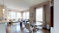This single-floor space showcases a seamlessly integrated open-concept layout that combines a living area and dining space into one cohesive environment. Rich, dark hardwood flooring extends throughout, providing a warm and elegant foundation that contrasts beautifully with the light, neutral-toned walls.

The dining area features a round glass-top table with a chrome base, surrounded by contemporary white chairs with sleek curved frames, lending the space a modern, sophisticated character. Nearby, large windows dressed with white horizontal blinds and floor-length silver-grey drapes flood the area with abundant natural light.

The adjacent living area continues the modern aesthetic with a white upholstered sofa accented by multicolored throw pillows, a glass coffee table, and a dark rectangular ottoman. A flush-mount ceiling light fixture adds a subtle decorative touch overhead. Additional windows along the living room wall further enhance the bright, airy atmosphere.

A framed piece of wall art serves as a tasteful accent between the two zones. Overall, the space presents a clean, contemporary design with a well-balanced combination of light tones, reflective surfaces, and warm wood elements.