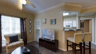 This single-floor space offers a cohesive layout where the living area transitions seamlessly into the kitchen. The living room features hardwood-style flooring, crown molding, and neutral warm-toned walls that create a welcoming atmosphere. A ceiling fan is centrally positioned, contributing to both comfort and airflow. Wall art and decorative mirrors add character to the space, while a glass-paneled door provides natural light and access to the exterior.

The kitchen is partially open to the living area, separated by a raised counter with bar seating. This peninsula-style counter features a dark granite surface, providing a functional space for casual dining. The kitchen is equipped with white cabinetry and appliances, including a microwave, and benefits from overhead lighting that keeps the area bright and practical. The bar stools at the counter offer an informal dining option that enhances the open-concept feel.

Overall, the space presents a functional and comfortable design with consistent finishes throughout, including crown molding and coordinated wood-tone flooring, giving the interior a polished and unified appearance.