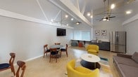 Located at 2858 W Ball Rd in Anaheim, California, this single-floor community lounge space offers a thoughtfully designed gathering environment with a distinctly modern aesthetic. The room is defined by its dramatic vaulted ceiling with exposed white structural beams, which lend an open, airy quality throughout the interior. Multiple ceiling fans and a striking brass sputnik chandelier provide both function and visual interest overhead.

The layout is organized into several distinct seating zones. A central lounge area features a plush gray sofa, vibrant yellow accent chairs, and a round coffee table set atop a colorful area rug, creating an inviting focal point. Adjacent to this is a casual dining area with a round table and mid-century style wooden chairs with navy upholstery. A built-in wall-mounted television serves this zone as well.

One wall is finished in a deep teal board-and-batten panel treatment, adding a bold architectural accent and grounding the space with rich color. Natural light filters in through large windows fitted with white blinds, and a set of glass French doors opens to an outdoor area beyond.

A compact kitchenette along one wall includes a stainless steel sink, gray cabinetry, and a full-size stainless steel refrigerator, making the space well-suited for events and gatherings. Polished concrete-style flooring ties the entire space together with a clean, contemporary finish.