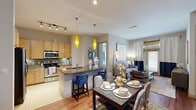 This single-floor residence offers a thoughtfully designed open-concept layout that seamlessly connects the kitchen, dining, and living areas. The kitchen is well-equipped with light maple cabinetry, stainless steel appliances including a refrigerator, microwave, and range, complemented by a granite countertop peninsula with seating for bar stools. Two striking yellow pendant lights hang above the peninsula, adding a bold design accent alongside a track lighting system overhead. The dining area accommodates a full dining table and benefits from hardwood flooring that continues into the living room. The living room is a comfortable retreat featuring hardwood floors, natural light from a window with layered curtains and wood blinds, and ample space for lounge seating. A dedicated dining nook with track lighting provides additional entertaining space. The two bedrooms are carpeted and well-proportioned, each featuring ceiling fixtures and windows with blinds that allow for natural light. The full bathroom includes a granite vanity with wood cabinetry, a tiled soaking tub with shower combination, and tasteful wall decor. Throughout the residence, neutral wall tones and consistent finishes create a cohesive and inviting atmosphere.