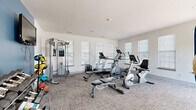 Located at 1001 Pinehurst Drive in Spring Hill, Tennessee, this dedicated fitness center offers a well-equipped exercise environment designed to support a wide range of workout routines. The single-level space features neutral-toned walls, with one side accented by a bold blue wall that adds a modern, energizing touch. Durable, textured carpet flooring covers the entire area, providing comfort and traction during workouts.

Natural light streams in generously through multiple windows fitted with white horizontal blinds, creating a bright and inviting atmosphere. The room is equipped with a comprehensive selection of fitness equipment, including a treadmill, elliptical trainer, upright and recumbent stationary bikes, and a multi-function cable weight machine with an adjustable bench. A full dumbbell rack offers a wide range of free weights, while a dedicated exercise ball storage stand holds an assortment of medicine and stability balls for functional training.

A wall-mounted flat-screen television is positioned for easy viewing during workouts. The space also includes a coat hook area near the entry and is marked with a clearly visible exit sign for safety. Access is provided through a glass-paneled door. The overall design is clean, functional, and well-organized, making this fitness center a practical and motivating space for daily exercise.