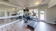 This single-level fitness center offers a comprehensive workout environment designed to accommodate a variety of exercise routines. The space is finished with durable wood-look flooring throughout and features crisp white walls accented by crown molding, giving the room a clean and polished appearance. Flush-mount and pendant light fixtures provide consistent overhead illumination, complemented by abundant natural light streaming through multiple large windows that frame outdoor views of surrounding greenery and a pool area. A glass-paneled door provides direct access to the outdoor space, reinforcing the connection between interior and exterior. A full-length wall mirror visually expands the room while allowing users to monitor their form during exercise. The cardio section is equipped with a treadmill and elliptical trainer, while a multi-station cable machine and adjustable benches serve the strength training area. A tiered dumbbell rack offers a range of free weights, and a wall-mounted medicine ball rack supports functional fitness training. A yoga mat is stored on the wall, providing a dedicated space for stretching and floor exercises. Ceiling fans and HVAC vents contribute to a comfortable climate throughout workouts.