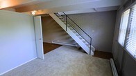 This two-story space showcases a blend of industrial and residential character, anchored by a prominent exposed brick wall that adds texture and warmth throughout the main living area. A sleek, open-riser staircase with a black metal railing serves as a central architectural feature, connecting the two floors while maintaining an open, airy feel. The main level is finished with neutral carpeting that extends across the living area, complemented by hardwood flooring accents that add visual contrast and depth. Exposed ceiling beams contribute to the raw, loft-like aesthetic of the space. A baseboard heating unit runs along the base of the brick wall, ensuring practical comfort. A ceiling fan provides additional climate control and light to the lower level. A door on the main floor leads to an adjacent room or closet, adding functional storage or additional living space. The overall ambiance is understated and versatile, with the combination of brick, open staircase, and mixed flooring materials creating a space that balances utilitarian design with an inviting atmosphere well-suited for a variety of uses.