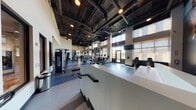 Located along John Muir Parkway in Hercules, California, this single-story fitness center offers a spacious, thoughtfully designed workout environment suited for a wide range of exercise routines. The facility is characterized by soaring ceilings with exposed ductwork and industrial-style black structural elements, lending the space a contemporary, open aesthetic. Recessed and pendant lighting throughout ensures consistent illumination across all areas.

Large floor-to-ceiling windows line one side of the facility, flooding the interior with natural light and providing views of the surrounding outdoor landscape. The cardio zone features a generous row of treadmills and stationary bikes positioned along the windowed wall, allowing users to exercise while enjoying outdoor views. Mounted flat-screen televisions are positioned throughout for entertainment.

The strength training area is equipped with a variety of cable machines, multi-station units, free weights, and benches, all arranged on durable rubber flooring in a speckled blue-grey finish. A dedicated open floor zone provides space for functional training and stretching. A vibrant athletic mural anchors one end of the facility, adding visual energy to the space. Motivational artwork and a water station are also present near the restroom corridor, enhancing the overall user experience.
