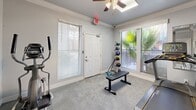 Located at 3131 Hayes Rd in Houston, Texas, this dedicated exercise room offers a practical and well-equipped fitness environment. The space features soft gray carpeting underfoot and crisp white crown molding along the ceiling, lending a clean and polished aesthetic. Large windows fitted with horizontal blinds allow natural light to filter in, providing views of lush tropical greenery outside, including a visible palm tree that adds a pleasant outdoor connection. A ceiling fan with an integrated light fixture keeps the room comfortable during workouts. The room is equipped with a Matrix treadmill, an elliptical machine, a weight bench, medicine balls on a vertical rack, and a CAP fitness mat, offering a well-rounded selection of cardio and strength training options. Vibrant abstract artwork decorates the walls, adding a touch of color and energy to the space. A posted fitness rules and regulations sign reflects the room's shared-use nature. An interior glass-panel door connects to an adjacent area, while a separate exterior door provides direct access to the outside. The overall ambiance is bright, functional, and inviting for daily exercise.