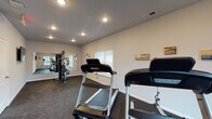 Located at 7407 Fegenbush Ln in Louisville, Kentucky, this single-floor fitness center is purpose-built to support a well-rounded workout routine. The space is laid out in a rectangular floor plan, providing ample room to accommodate multiple users and equipment simultaneously without feeling cramped.

The flooring throughout consists of durable, dark rubber gym matting designed to absorb impact and reduce noise — a practical choice well-suited to high-traffic exercise environments. Neutral gray walls create a clean, focused atmosphere, while recessed ceiling lighting ensures the space is evenly and brightly lit. Emergency exit signage is clearly positioned, reflecting a commitment to safety.

Equipment includes multiple Spirit-brand treadmills positioned to allow comfortable use, along with a multi-station cable and strength machine. A large wall-mounted mirror on one end visually expands the space and provides form-checking capability during workouts. A flat-screen television is mounted on the wall, offering entertainment during cardio sessions.

Several windows fitted with white horizontal blinds allow natural light to enter and provide views of the surrounding exterior. Framed artwork and small decorative pieces on the walls contribute a welcoming touch. A keyed entry door with a sidelight window provides private, controlled access to the facility.