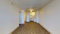 This single-floor unit offers a straightforward and functional layout designed to maximize usable space. The main living area is finished with warm wood-style laminate flooring that runs throughout, providing a cohesive and clean aesthetic. A ceiling fan with integrated lighting serves as the central fixture, offering both comfort and illumination to the open space. Crisp white walls and a textured ceiling contribute to a bright, neutral atmosphere throughout.

The kitchen is positioned at the rear of the main living area and is open to the space, creating a connected feel. It features white upper and lower cabinetry, a white refrigerator, a white range, and a standard sink with countertop workspace. The cabinetry is well-organized, offering practical storage solutions. A doorway leading to an additional room or corridor is visible to the left, suggesting further living accommodations accessible from the main space.

The exposed brick-style wall detail along the side adds a subtle textural element to the otherwise smooth interior. The open floor plan and seamless transition between the living and kitchen areas make this a practical and easy-to-navigate space. Overall, the unit presents a clean, move-in-ready environment with functional finishes throughout.