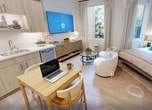 This 330-square-foot single-floor studio is designed to maximize functionality without sacrificing style. The open-plan living and sleeping area is bathed in natural light through large windows dressed with floor-length neutral curtains, offering views of lush outdoor greenery. Light oak-toned hardwood flooring runs throughout, unifying the space with warmth and continuity.

The kitchen is a standout feature, outfitted with light wood cabinetry, white quartz countertops, stainless steel appliances including a bottom-freezer refrigerator, built-in dishwasher, an induction cooktop, and a built-in microwave. A compact dining table with two chairs provides a flexible workspace or dining area within the kitchen zone.

The living and sleeping area offers generous space for a full-size bed alongside a cozy seating arrangement, complete with a glass coffee table and rounded accent chairs. Framed black-and-white artwork adds a curated, sophisticated touch to the neutral walls.

The bathroom features a walk-in glass-enclosed shower with a rainfall showerhead, a light wood vanity with a white countertop, built-in open shelving for toiletries, and wood-look tile flooring. A woven area rug adds texture and comfort underfoot. Clean lines and a neutral palette give the entire unit a calm, modern ambiance.