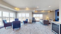 Located on Bear Path in Byron, Minnesota, this community room provides a well-appointed, single-floor shared space designed for comfortable gathering and socializing. The open layout is anchored by patterned, neutral-toned carpeting that extends throughout, complementing the warm, neutral wall tones and recessed lighting overhead. Tray ceiling details add architectural interest and a sense of depth to the space.

Multiple seating zones define the room's versatile layout. A lounge area features plush blue accent chairs arranged around a round coffee table, ideal for casual conversation. A separate area offers a sofa with decorative pillows alongside a bar-height bistro table with chairs, providing options for both relaxed and upright seating. A small beverage or kitchenette unit is visible along one wall, adding functional convenience.

Large windows along multiple walls flood the interior with natural light and offer views of the surrounding Byron neighborhood. Wall-mounted artwork and carefully selected accent pieces, including a leather bench and side tables near the entry, contribute to the room's polished, welcoming ambiance. A restroom is accessible directly from the space. Overall, the community room balances practicality with a modern, comfortable aesthetic suited to a variety of social occasions.