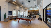This contemporary single-floor clubhouse is designed to offer a well-rounded amenity experience, combining entertainment, relaxation, and community gathering in one cohesive space. The welcoming entrance foyer sets the tone with soaring ceilings, large-format tile flooring laid in a herringbone pattern, bold abstract artwork, warm wood ceiling accents, and floor-to-ceiling glass doors that flood the space with natural light.

The main gathering area is anchored by a full-size billiard table and benefits from impressively high ceilings with recessed lighting and ceiling fans. A full kitchen sits adjacent, featuring light wood cabinetry, a bold geometric tile backsplash, stainless steel appliances, a spacious quartz island with bar seating, open shelving, and pendant lighting — ideal for entertaining or community events.

A dedicated media lounge offers plush seating, wall-mounted displays, and built-in cabinetry, creating a comfortable space for screen viewing. A tech-forward workstation area with high-top seating and mounted monitors provides a functional workspace within the clubhouse.

Floor-to-ceiling sliding glass doors open directly onto a covered outdoor patio furnished with modern lounge seating, overlooking a landscaped pool area. Throughout the space, a consistent palette of neutral tones, natural wood elements, and clean architectural lines creates a polished yet inviting atmosphere.
