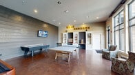 This energetic game room offers a well-appointed recreational space designed for entertainment and social interaction. The open, single-floor layout provides generous room for multiple activities simultaneously, centered around a ping-pong table, an air hockey table, and a billiards table — making it a versatile hub for competitive play and leisure.

The interior design blends modern and casual aesthetics, featuring a prominent shiplap accent wall, polished concrete flooring in warm brown tones, and recessed lighting throughout. A custom built-in shelving unit lines one wall, displaying board games, classic tabletop games such as Connect Four and Jenga, and decorative items. Bold oversized Scrabble-tile lettering spelling "LIVE VARSITY" adds a playful, spirited focal point above the shelving.

Wall-mounted flat-screen televisions are positioned for easy viewing from multiple vantage points within the room. A seating area with patterned wingback chairs is positioned near floor-to-ceiling windows, which flood the space with natural light and offer views of an outdoor pool and surrounding area — creating an inviting spot to relax between games. Decorative artwork adds character throughout. The room is accessible via multiple entry points, ensuring a welcoming and functional flow for all users.