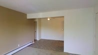 This single-floor space presents a simple, open layout characterized by neutral-toned walls and wall-to-wall carpeting throughout. The main living area flows into an adjoining room defined by a structural soffit overhead, creating a subtle visual separation between spaces without fully enclosing them. A ceiling fan with a light fixture is mounted in the adjoining area, providing both air circulation and ambient lighting. Along one wall, a baseboard heating unit ensures efficient climate control throughout the colder months. A doorway visible along the side wall leads to an additional room or closet space, suggesting practical storage or private quarters nearby. The walls are painted in a soft, warm beige tone, contributing to a neutral and understated ambiance. The space is modest in scale but benefits from its open-plan design, which allows for flexible furniture arrangement. Overall, the interior presents a clean, utilitarian aesthetic well-suited for comfortable everyday living.