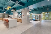 Located in the heart of Midtown Manhattan, this expansive single-floor commercial space at 419 W 34th Street offers a thoughtfully designed environment that blends modern aesthetics with functional amenities. The interior is immediately striking, featuring bold teal-painted exposed ceilings with structural beams and ductwork that lend an industrial-chic character, contrasted by clean large-format light grey tile flooring throughout.

The open main area flows seamlessly between a variety of zones. A spacious dining and kitchen area serves as a central hub, equipped with a long communal bar-height counter lined with vibrant orange seating, wood-accented cabinetry, built-in appliances, and patterned tile backsplashes. Pendant lighting in mixed styles — globe, cylindrical, and sculptural — adds visual interest overhead. Vending machines and casual seating clusters provide convenience for daily use.

Arched passageways connect the main space to additional seating rooms, including a bright dining nook with sage-green walls, natural light from large multi-pane windows, and a modern dining table with white and green-framed chairs. A service counter and reception-style area anchor one end of the floor.

Outside, a landscaped courtyard with brick paving, mature trees, greenery, and casual seating offers a tranquil retreat, complemented by a secondary outdoor walkway along the building's historic brick façade.