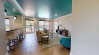 This single-floor community space makes an immediate impression with its signature teal-painted textured ceilings, accented by crisp white crown molding throughout. Large-format stone tile flooring anchors the open layout, lending a warm, grounded aesthetic that flows seamlessly from one area to the next.

The main lounge area is generously sized, furnished with neutral sectional sofas, accent chairs in coordinating teal, and glass-top coffee tables set atop a multicolored area rug. Recessed lighting and an abundance of natural light streaming through sliding glass doors and large windows create a bright, welcoming atmosphere. These doors open to a fenced outdoor area with direct views of a swimming pool.

Adjacent to the lounge, a dining and bar area features a long counter-height bar with white upholstered stools, illuminated by a trio of striking starburst pendant lights. A separate dining table with modern chrome-legged chairs sits nearby, offering additional seating beneath its own statement chandelier. Structural columns define the zones without interrupting the open feel of the space.

A kitchenette at the rear of the space is equipped with dark cabinetry, granite countertops, a stainless steel refrigerator, and a sink — ideal for hosting and events. Decorative framed artwork is displayed throughout, adding color and personality to the overall design.