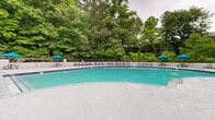 This outdoor amenity space centers around a generously sized swimming pool with clear turquoise water, framed by a wide, smooth concrete deck designed for both relaxation and socializing. The pool's curved shape adds visual interest and a resort-like quality to the setting, with stainless steel entry rails providing safe and convenient access.

Encircling the pool deck is an extensive lounge area furnished with multiple gray adjustable chaise lounges, ideal for sunbathing or unwinding. Bright blue market umbrellas are positioned throughout, offering shaded retreats, while picnic-style tables provide additional gathering spots. A raised lounge platform features stylish pod-shaped wicker chairs and a stone-accented outdoor bar or grill station beneath a white pergola structure, creating a distinct entertainment zone.

A charming poolside building with a gabled roof and white pergola entry serves as a pool house or utility structure, surrounded by well-maintained landscaping including manicured shrubs, mulched garden beds, and a mature shade tree at the entrance. The entire area is enclosed by a black metal fence, with a dense canopy of tall trees providing a lush, natural backdrop that enhances the sense of privacy and tranquility throughout the space.