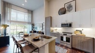 This single-floor residence is defined by its dramatic soaring ceilings and open-concept layout, creating an expansive, airy atmosphere throughout. The living area is anchored by floor-to-ceiling sliding glass doors that flood the space with natural light and provide direct access to a private balcony overlooking lush green surroundings. Sheer linen drapes frame the oversized windows, softening the abundant daylight while maintaining the sense of openness.

The kitchen is thoughtfully designed with two-tone cabinetry—crisp white upper cabinets paired with warm gray lower cabinets—complemented by granite countertops, a white subway tile backsplash, and a full suite of stainless steel appliances. A kitchen island with an undermount sink and bar seating serves as both a functional workspace and a casual dining area. Rich hardwood flooring flows seamlessly from the kitchen into the living space.

The bedroom offers a refined retreat with plush carpeting, darkening drapes, and contemporary décor. An adjacent bathroom features a granite-topped vanity with warm wood cabinetry and a large mirror, opening directly to a spacious walk-in closet with open shelving for organized storage. Every detail throughout reflects a cohesive, modern aesthetic.