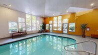 Located in Kent, Washington, this indoor aquatic facility offers a generously sized swimming pool as its centerpiece, surrounded by a wide pool deck with ample room for seating and relaxation. The space is defined by soaring vaulted ceilings that lend an open, airy feel, while large floor-to-ceiling windows along multiple walls flood the interior with natural light and frame views of the lush, tree-lined surroundings outside.

The pool area features stainless steel entry rails, safety equipment, and informational signage, reflecting a well-maintained and safety-conscious environment. Seating benches and potted bamboo plants are thoughtfully placed throughout the deck, adding a touch of warmth and greenery to the space.

A standout architectural feature is the circular hot tub alcove, enclosed by a curved glass block wall that diffuses light beautifully and creates a private, spa-like atmosphere. A raised platform area with a bold blue accent wall and decorative plantings provides additional visual interest and separation within the facility.

The dual-tone color scheme — warm amber-yellow accent walls paired with crisp white walls and ceilings — gives the space an inviting and energetic character. The smooth concrete flooring throughout is both practical and clean in aesthetic, completing this well-appointed indoor aquatic retreat.